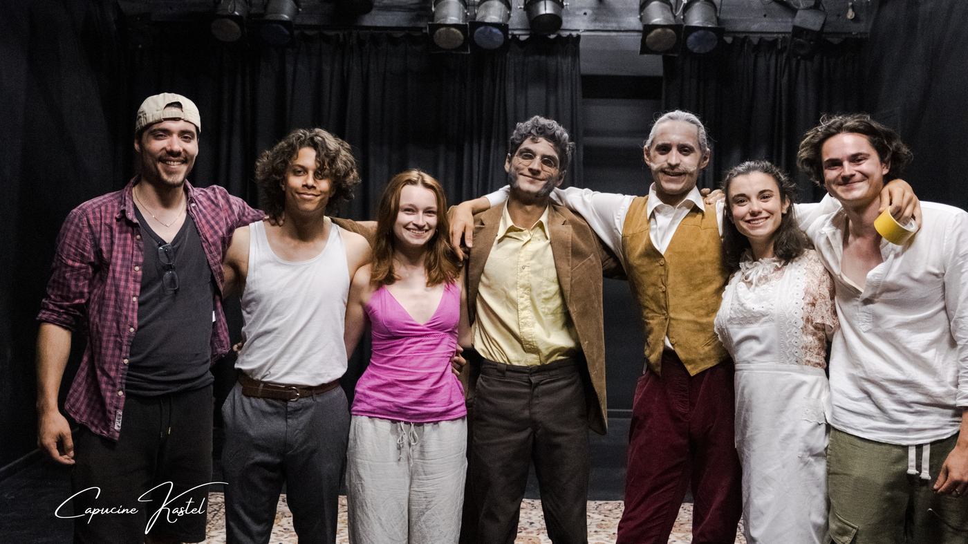 Sept membres de la compagnie, bras dessus bras dessous, sur un plateau de théâtre.